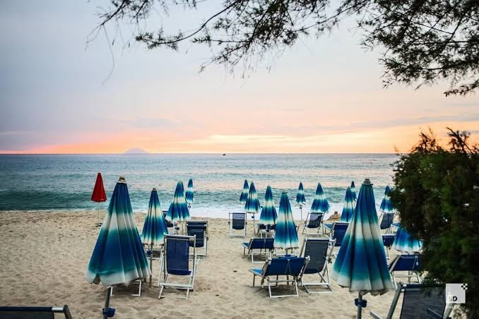 Tonicello Resort & Spa privātā pludmale Kalabrijā ar zili svītrainiem saulessargiem un zviļņiem smiltīs saulrieta laikā pie Tirēnu jūras