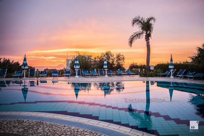 Panorāmas skats uz Tonicello Resort & Spa baseinu un jūras horizontu saulrietā — atpūtas krēsli un saulessargi piekrastē
