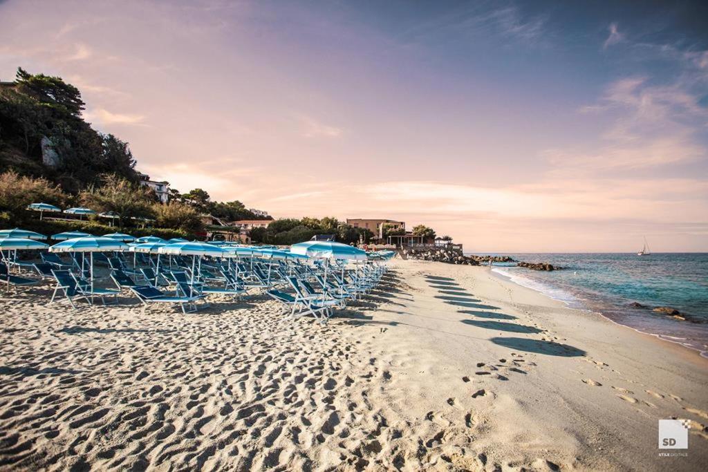 Skats uz Tonicello Resort & Spa pludmales zonu un apkārtnes pakalniem — smilšu krasts ar daudziem saulessargiem un atpūtas krēsliem
