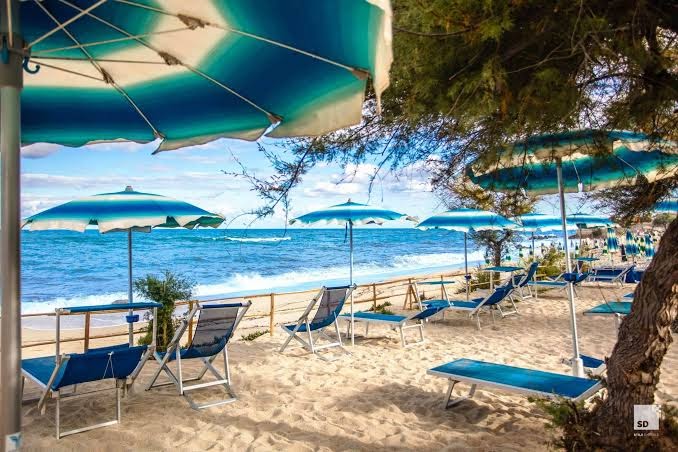 Tonicello Resort & Spa pludmales atpūtas zona zem kokiem — zili balti saulessargi un krēsli smiltīs pie pašas jūras