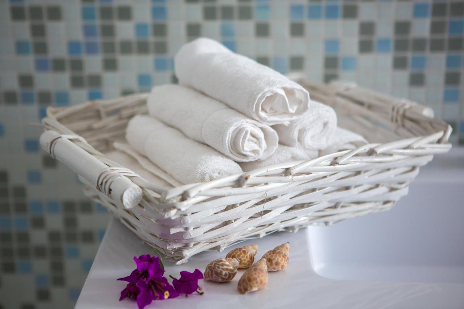Mīksti balti dvieļi pītā grozā Galia Luxury Resort vannas istabā, dekorēti ar gliemežvākiem un violetiem ziediem uz mozaīkas flīžu fona