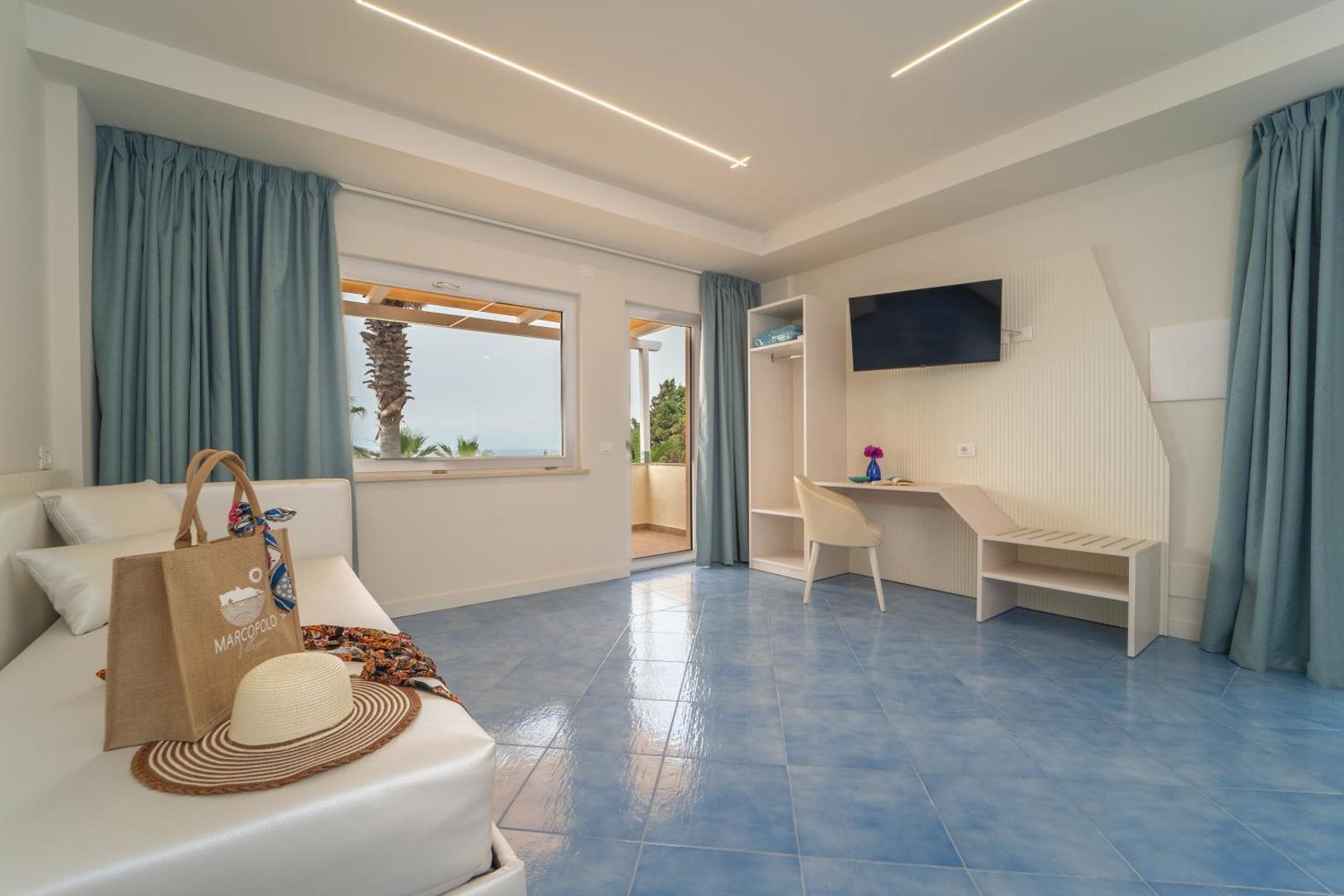 Villagio Marco Polo viesnīcas numurs ar gaiši zilām flīzēm, dīvānu, rakstāmgaldu un skatu uz palmu piekrastē Kalabrijā