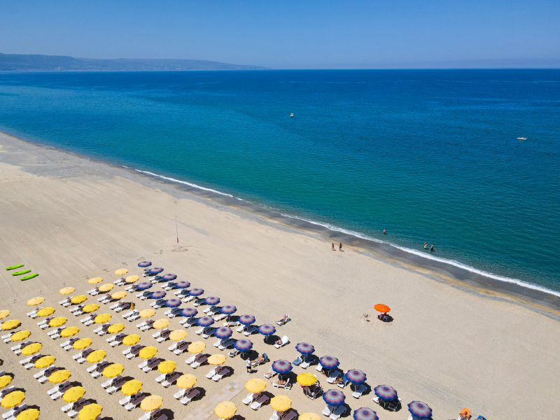 Futura Club La Praya plašā smilšu pludmale Kalabrijā ar dzelteniem un ziliem saulessargiem pie dzidri zilas jūras