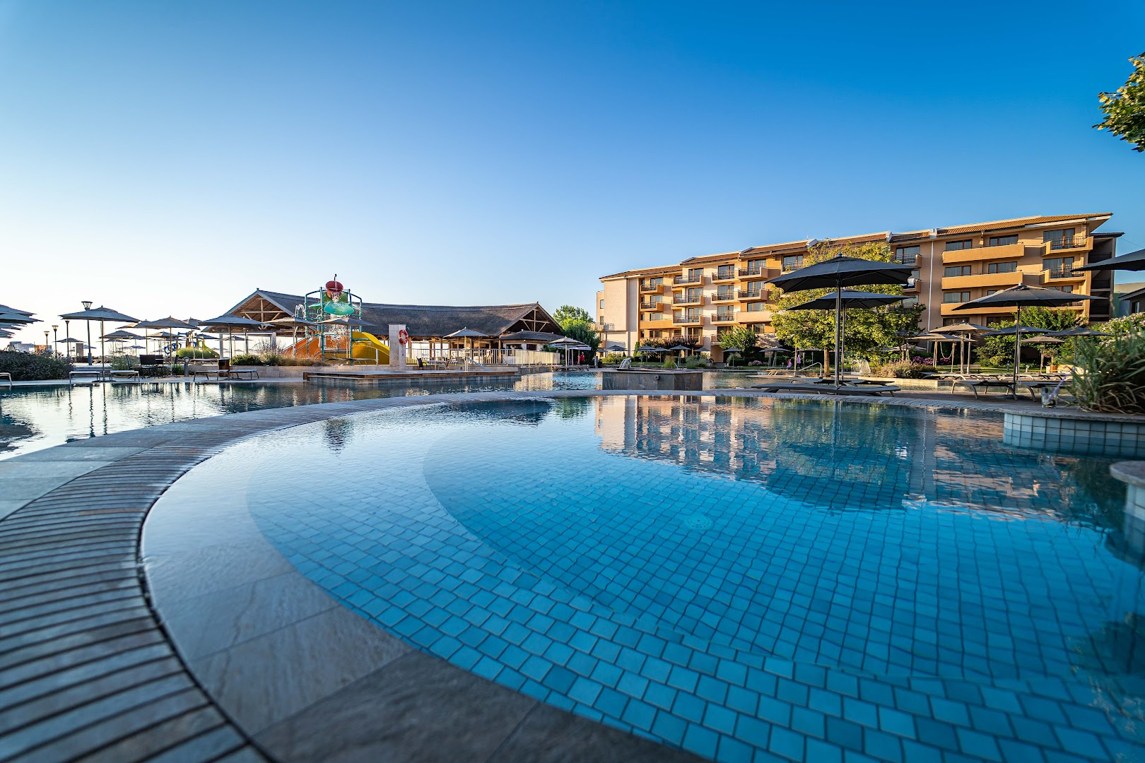 HVD Club Hotel Miramar bērnu baseins ar slidkalniņiem un galvenā ēka fonā Obzorā — saulains skats uz kūrorta zonu