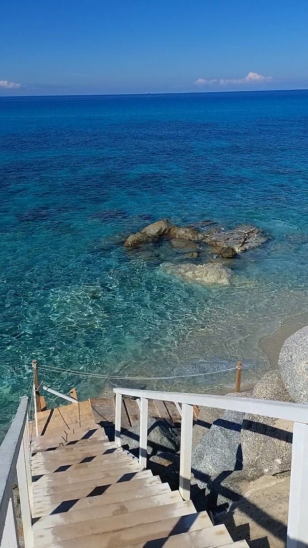 Skats no koka kāpnēm uz kristāldzidru jūras ūdeni un akmeņaino piekrasti Midenza Seaside & Nature Retreat teritorijā