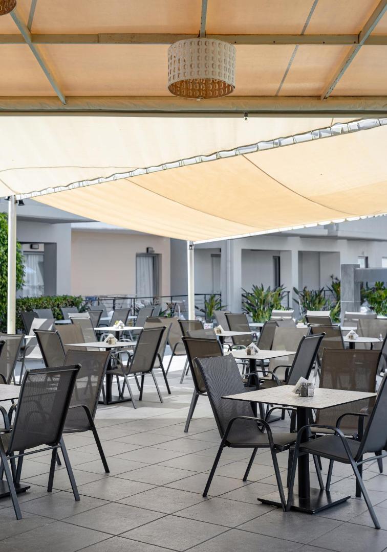Sunrise Village Hotel āra terase ar galdiņiem zem nojumes Krētas saulē — atpūtas zona maltītēm svaigā gaisā