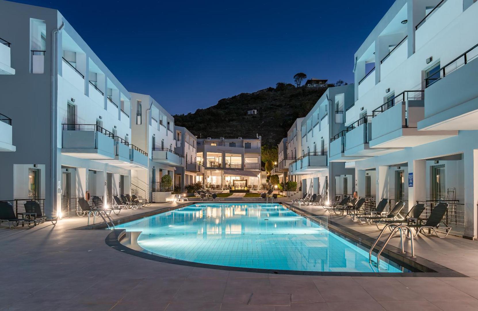 Sunrise Village Hotel izgaismots āra baseins vakarā starp viesnīcas ēkām uz kalna fona Krētā