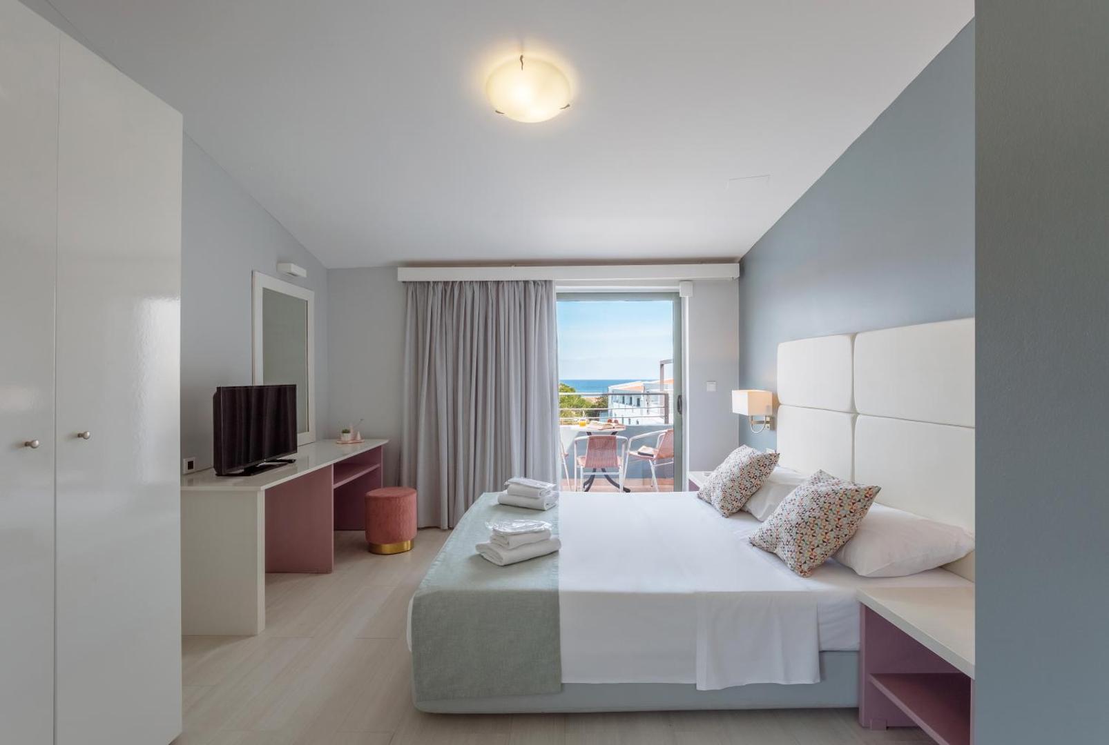 Sunrise Village Hotel gaišs divvietīgs numurs ar balkonu, no kura paveras skats uz jūru, modernām mēbelēm un rozā akcentiem