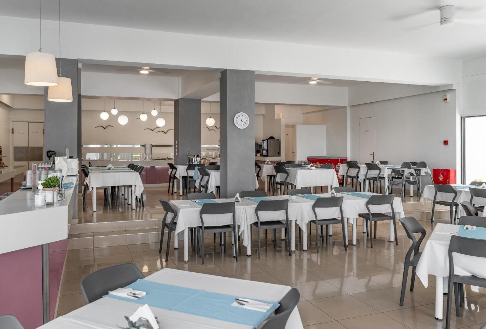 Sunrise Village Hotel galvenais restorāns ar klātiem galdiem un gaišu interjeru Krētā — plaša ēdamzāle ar modernām mēbelēm