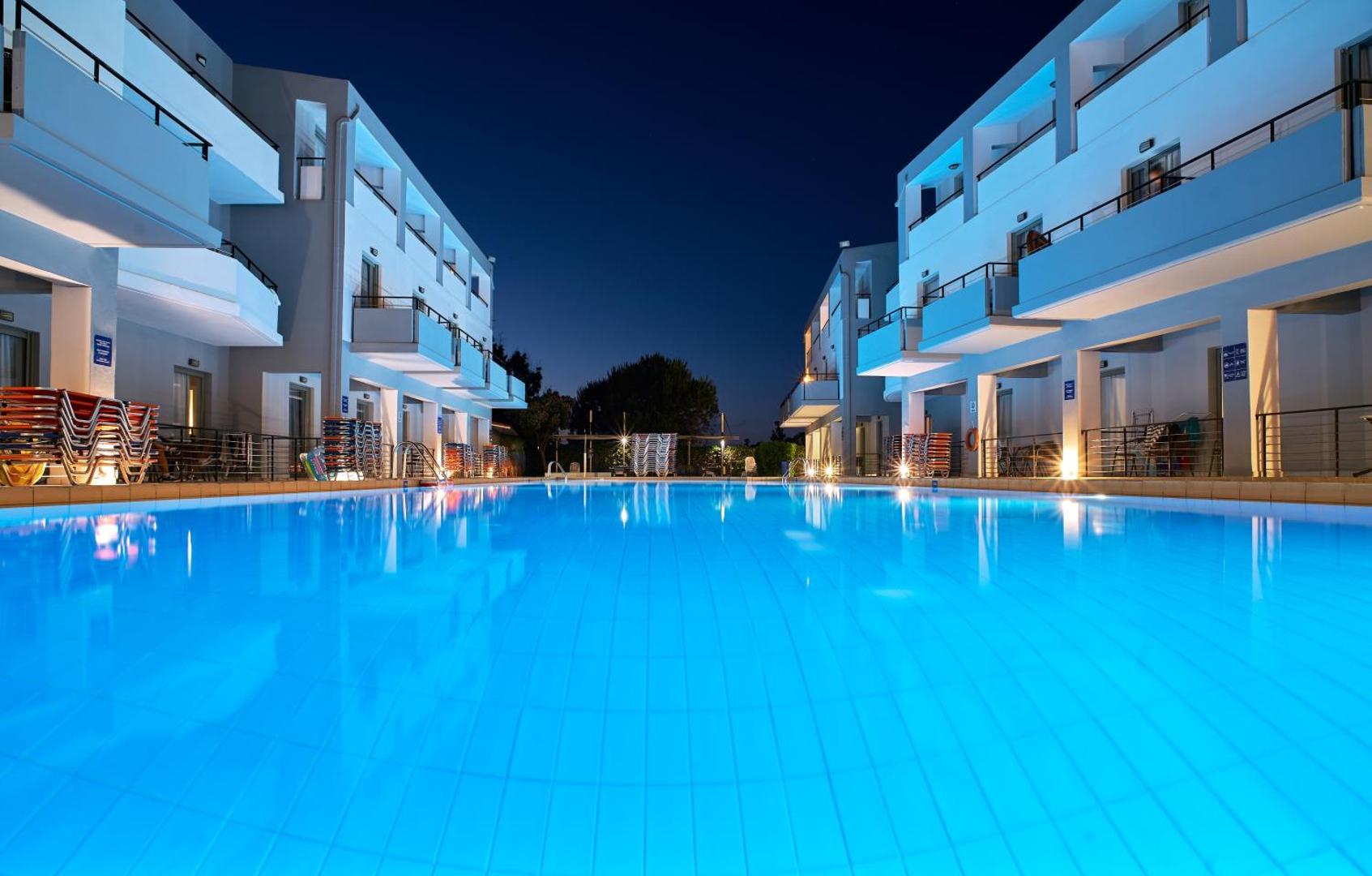 Sunrise Village Hotel baseina tuvplāns vakarā ar zilu apgaismojumu un viesnīcas korpusu atspulgu ūdenī