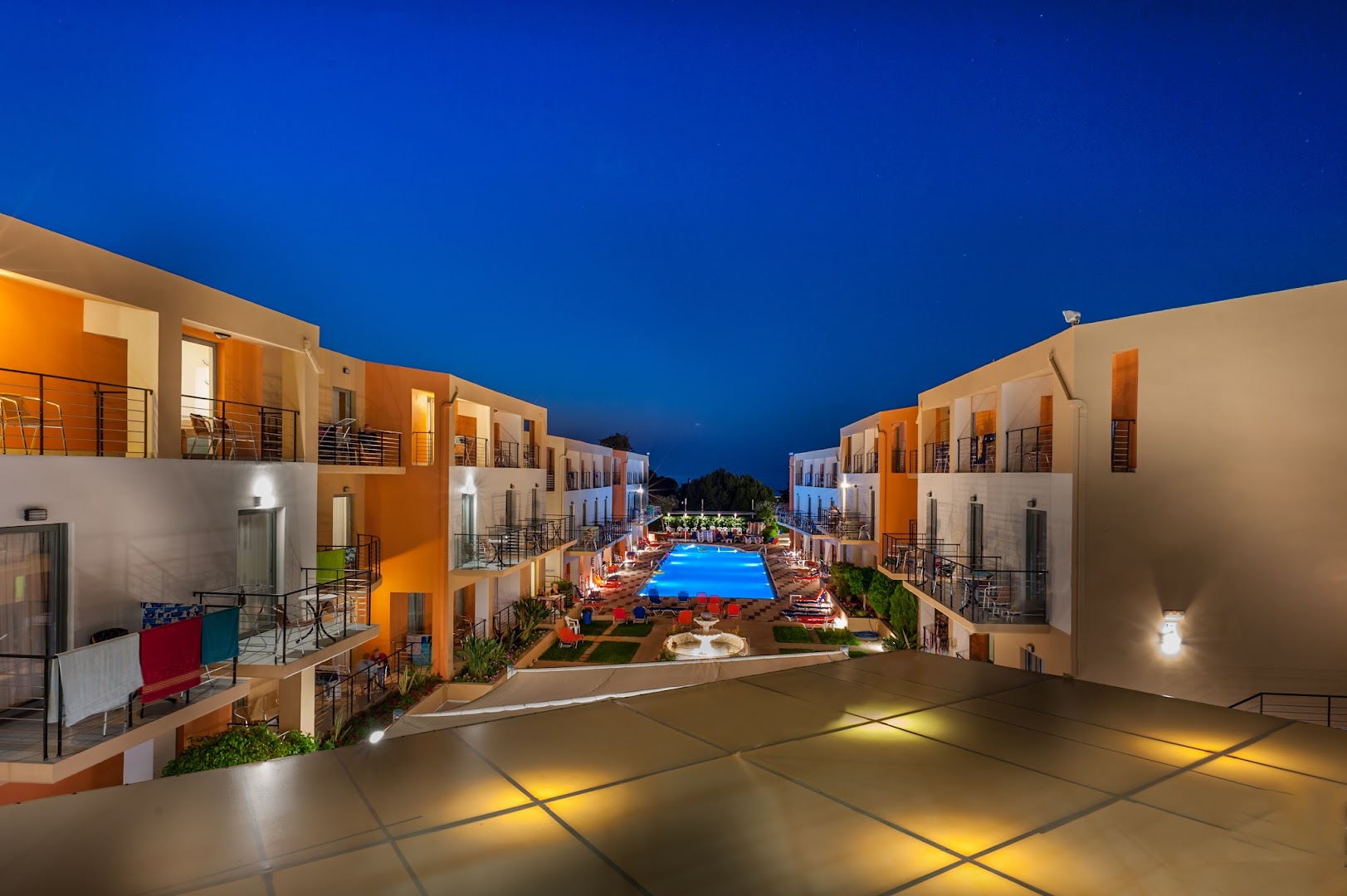 Sunrise Village Hotel iekšpagalms naktī ar izgaismotu baseinu un numuru balkoniem siltos toņos Krētas kūrortā