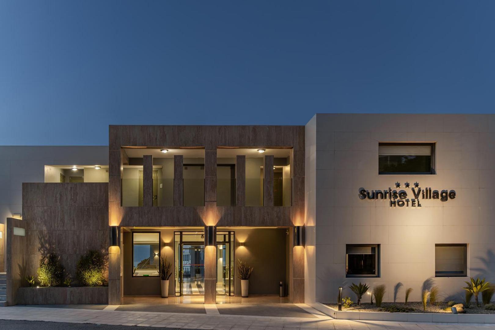 Sunrise Village Hotel modernā ieeja un fasāde vakara apgaismojumā Krētā — eleganta arhitektūra ar siltu gaismu un akmens apdari