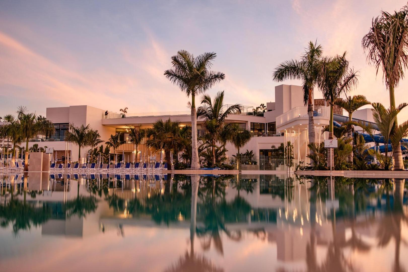 Servatur Puerto Azul Hotel galvenā ēka un baseins saulrieta laikā Grankanārijā — palmas un viesnīcas fasādes atspulgs ūdenī