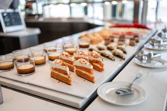 Servatur Puerto Azul Hotel desertu bufete Grankanārijā — burkānu kūkas gabaliņi un krēma deserti glāzītēs uz balta marmora virsmas
