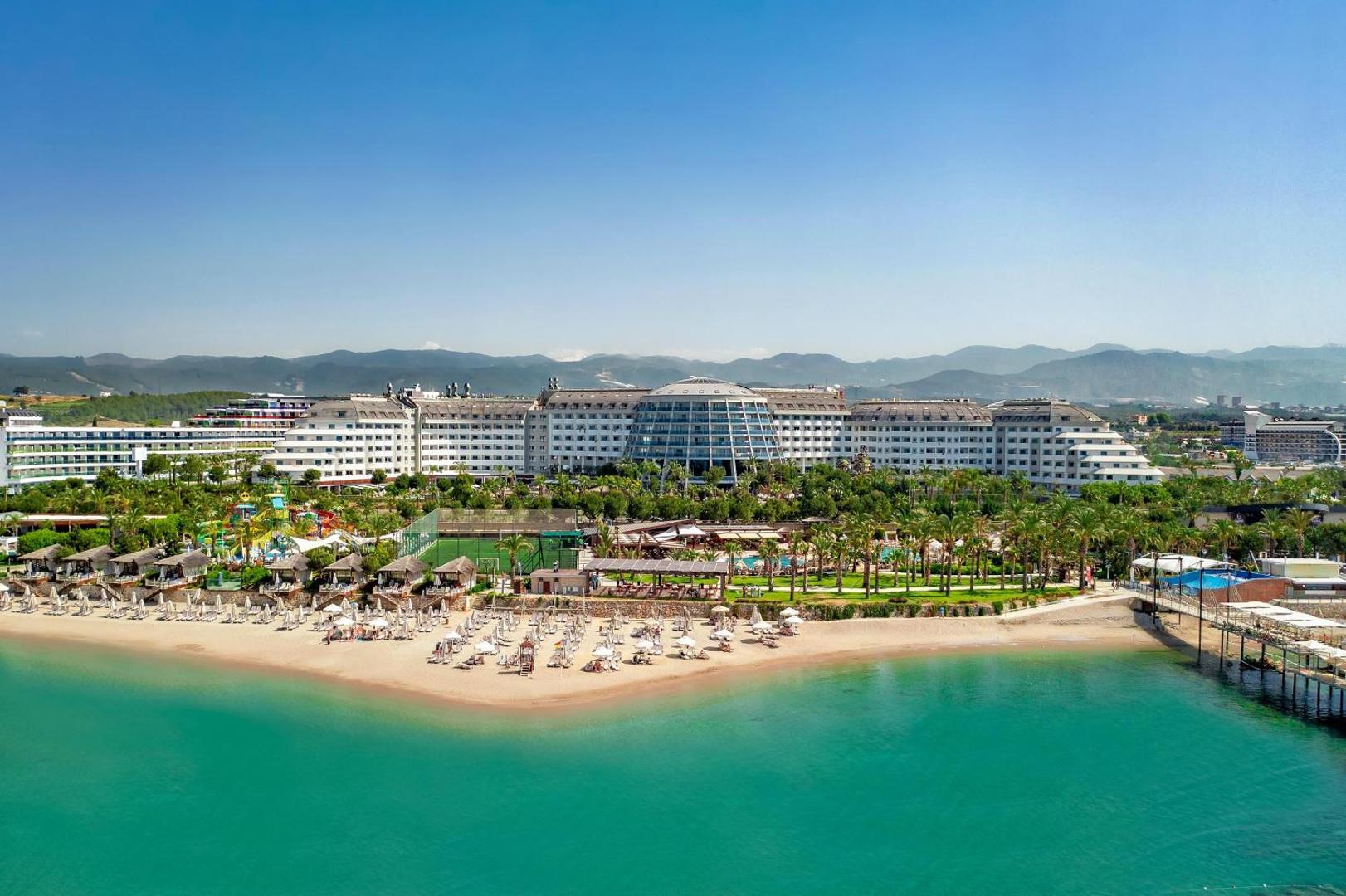 Long Beach Resort panorāmas skats no jūras puses Alānijā — plaša smilšu pludmale, viesnīcas galvenā ēka un kalni fonā