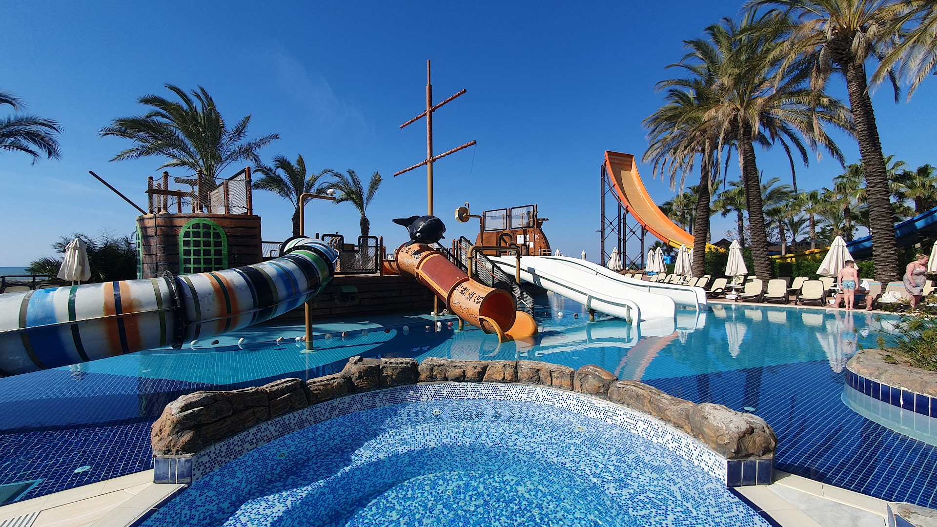 Long Beach Resort bērnu baseins ar pirātu kuģa tematikas slidkalniņiem Alanjā, Turcijā — sekls baseins un atpūtas zona ar palmām