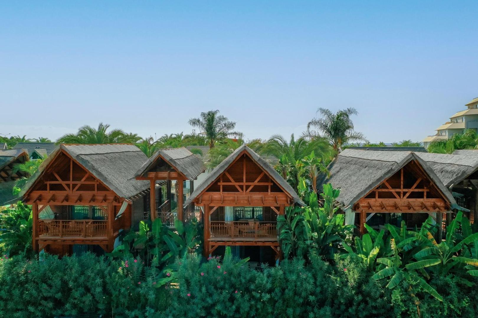 Long Beach Resort divstāvu koka bungalo ar niedru jumtiem un balkoniem starp zaļiem augiem Alānijā