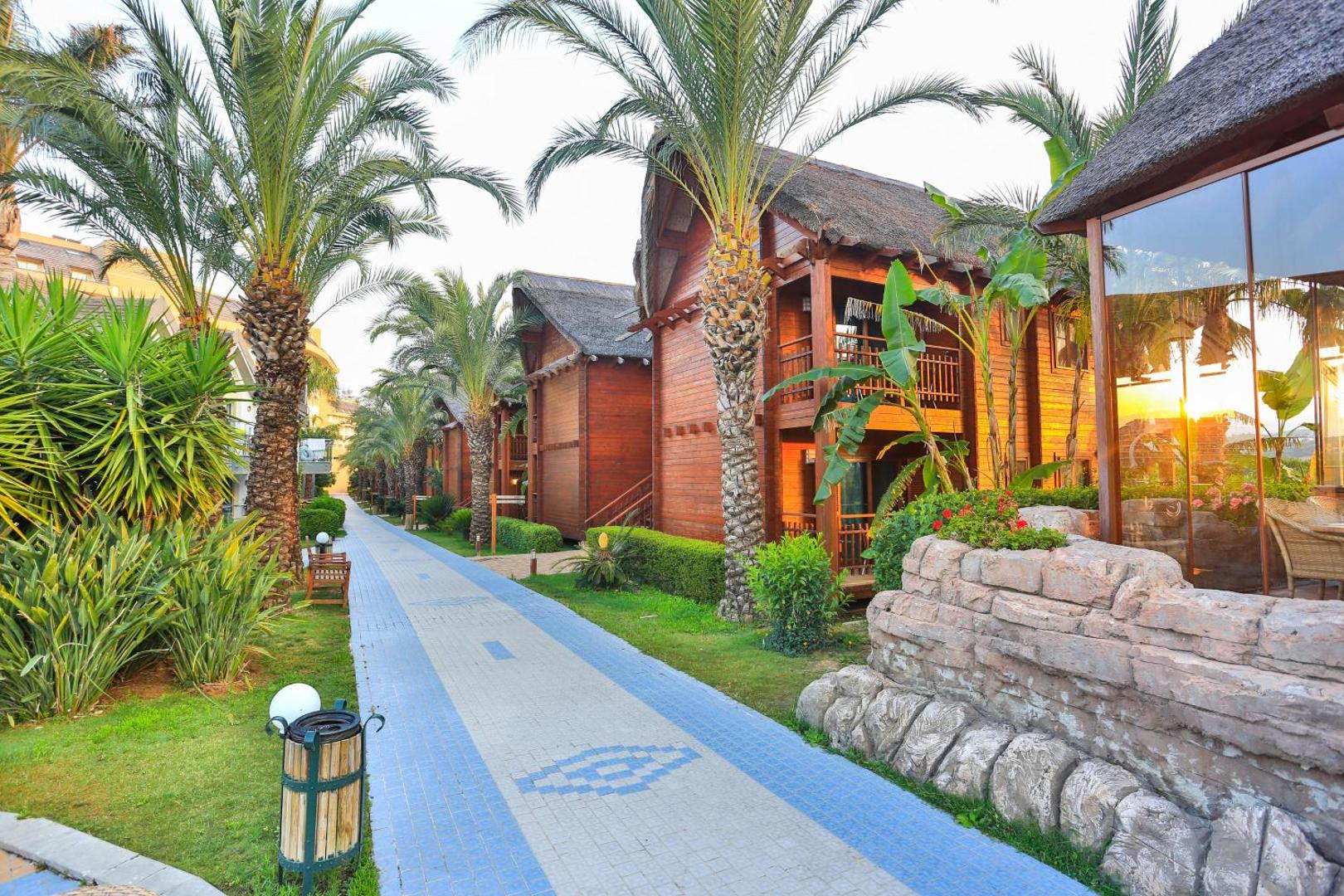 Long Beach Resort koka bungalo rinda ar palmu aleju un flīzētu gājēju celiņu saulrietā Alānijas reģionā