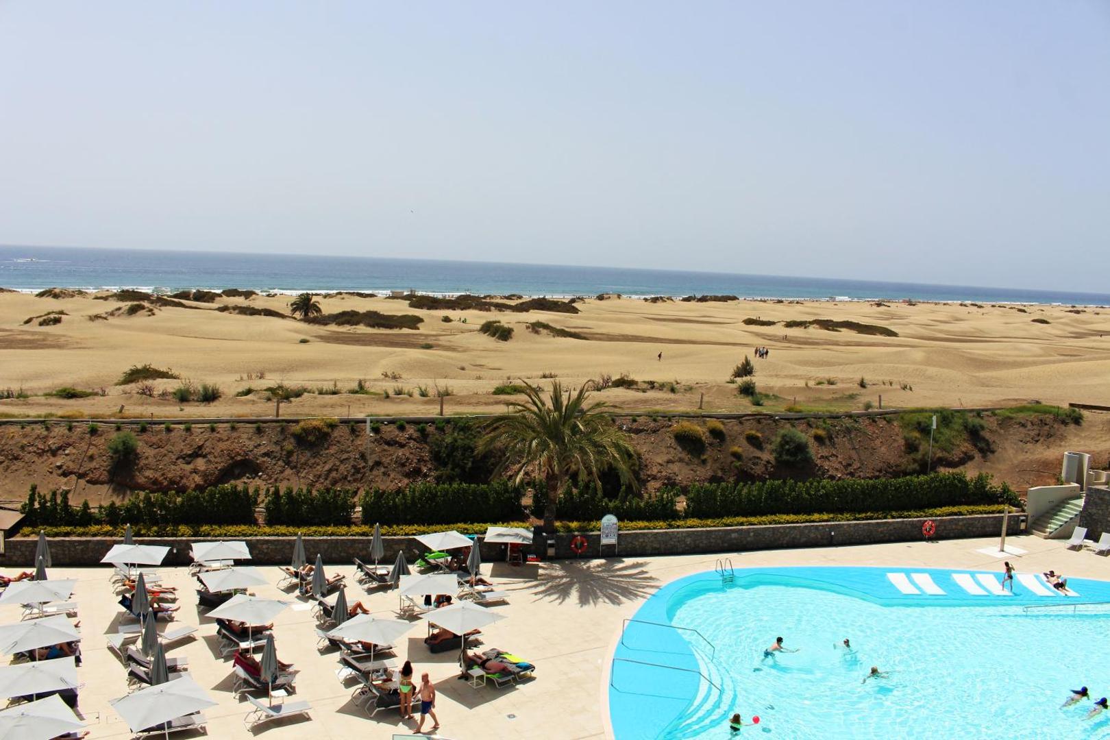 Santa Mónica Suites Hotel atpūtas zona pie baseina ar pelēkiem saulessargiem un skatu uz Maspalomas smilšu kāpām Grankanārijā
