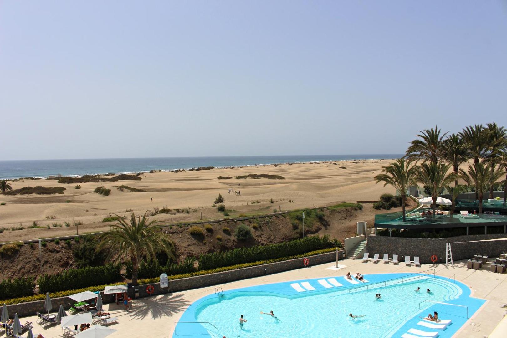 Skats no Santa Mónica Suites Hotel terases uz Maspalomas kāpām un okeānu — baseina zona ar atpūtas krēsliem un palmām