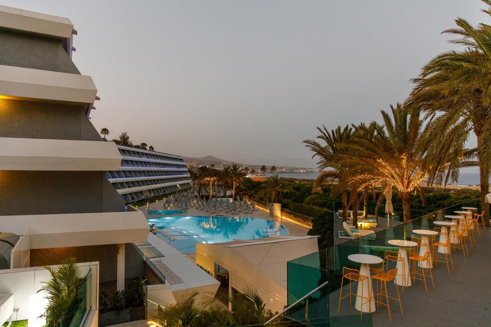 Santa Mónica Suites Hotel skats no augšas uz baseinu, palmu dārzu un viesnīcas korpusu saulrieta laikā Grankanārijā