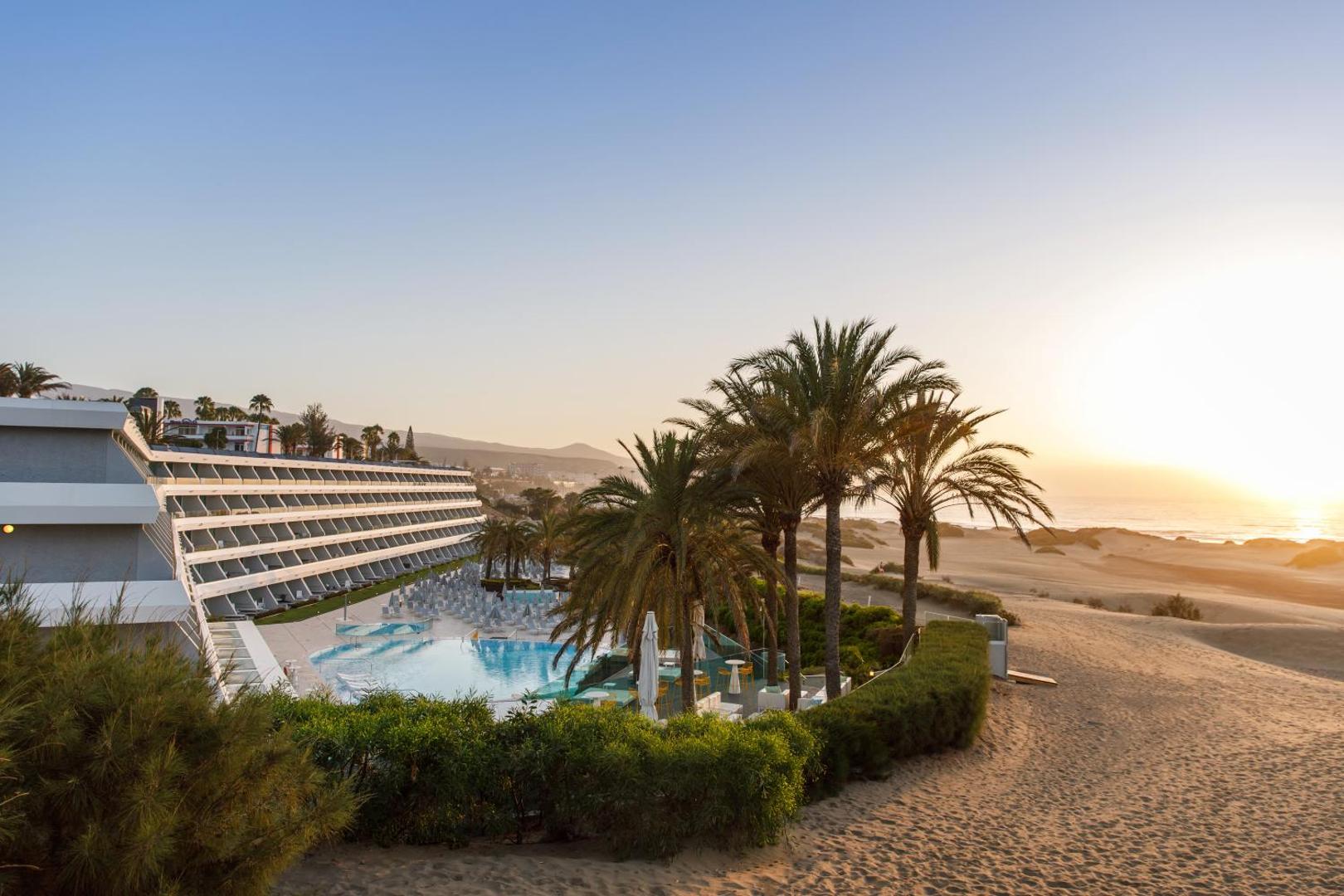 Santa Mónica Suites Hotel skats no augšas uz āra baseinu, palmām un Maspalomas kāpām saulrietā Grankanārijā