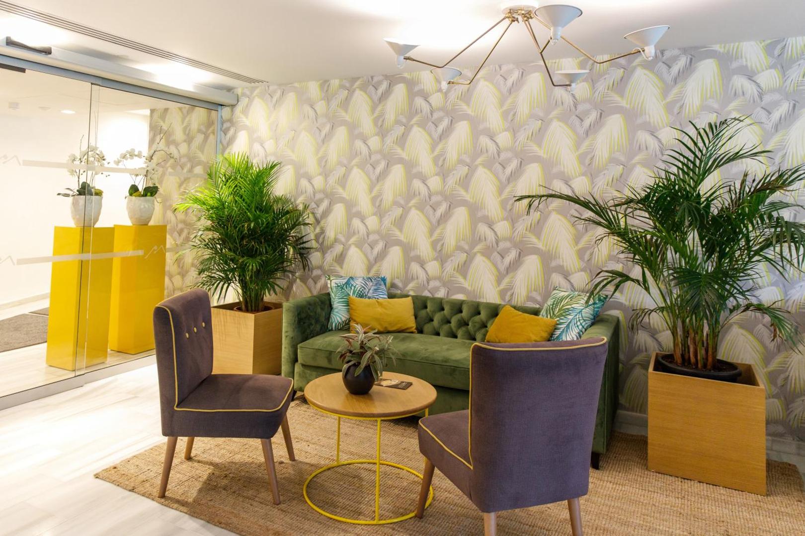 Santa Mónica Suites Hotel atpūtas zona ar zaļu dīvānu, violetiem krēsliem un tropu raksta tapetēm — stilīgs interjers ar telpaugiem