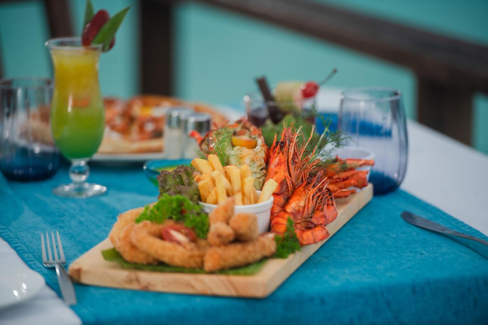 Makunudu Island restorāna jūras velšu plate ar garnelēm un frī kartupeļiem uz koka dēlīša, fonā eksotisks kokteilis Maldīvu salu pludmalē