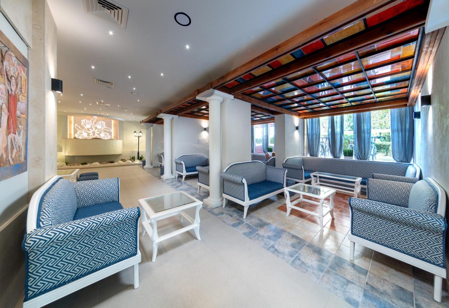 Sentido Bellevue Beach viesnīcas vestibils ar ziliem dīvāniem, baltiem galdiņiem un krāsainu vitrāžu griestu dekoru Saulainajā krastā, Bulgārijā