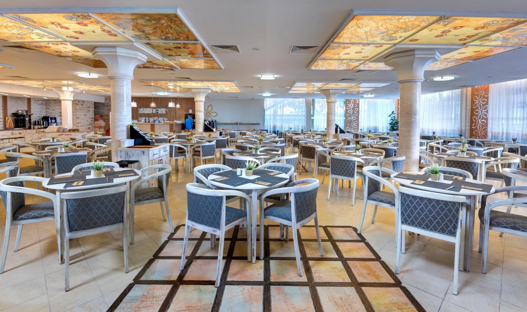 Viesnīcas Sentido Bellevue Beach plašā restorāna zāle ar daudziem galdiem, kolonnām un bāra zonu fonā