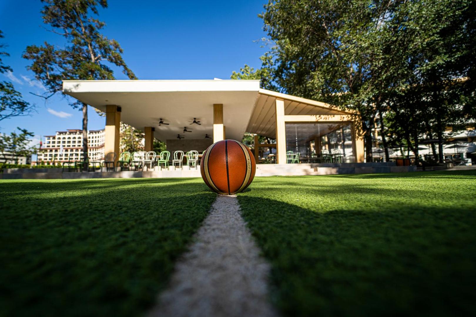 Basketbola bumba uz zaļā laukuma HVD Club Bor viesnīcas teritorijā — fonā redzama āra terase un koki