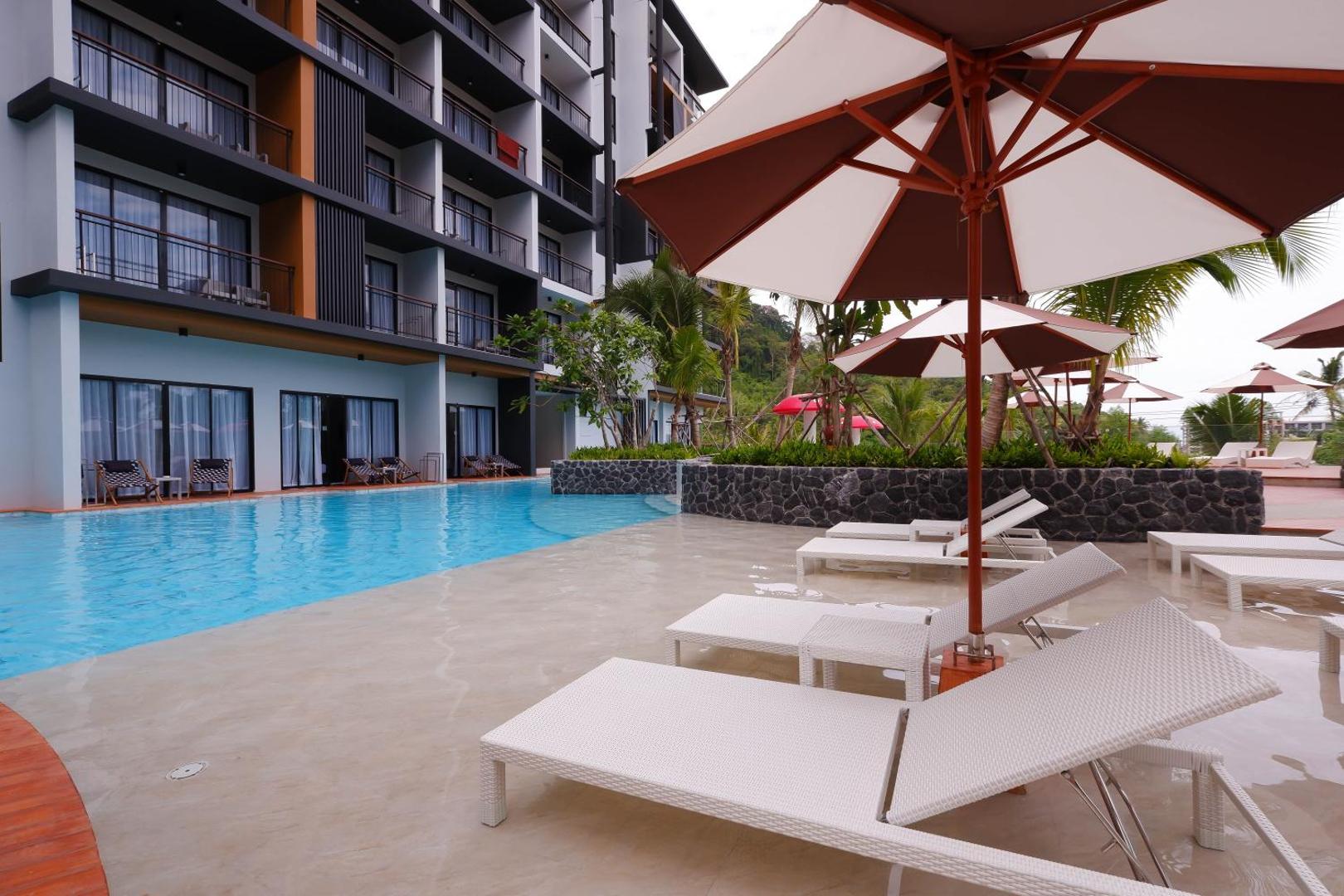 Sea Seeker Krabi Resort modernā fasāde un baseina zona ar baltiem zviļņiem un svītrainiem saulessargiem Ao Nangas piekrastē