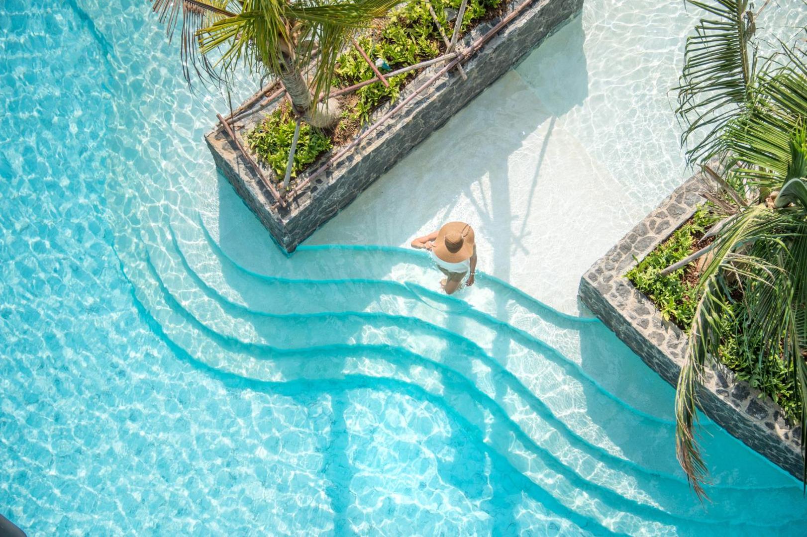 Viesis kāpj Sea Seeker Krabi Resort dzidrajā baseinā pa viļņveida pakāpieniem — saulains skats no augšas ar palmām un tirkīzzilu ūdeni