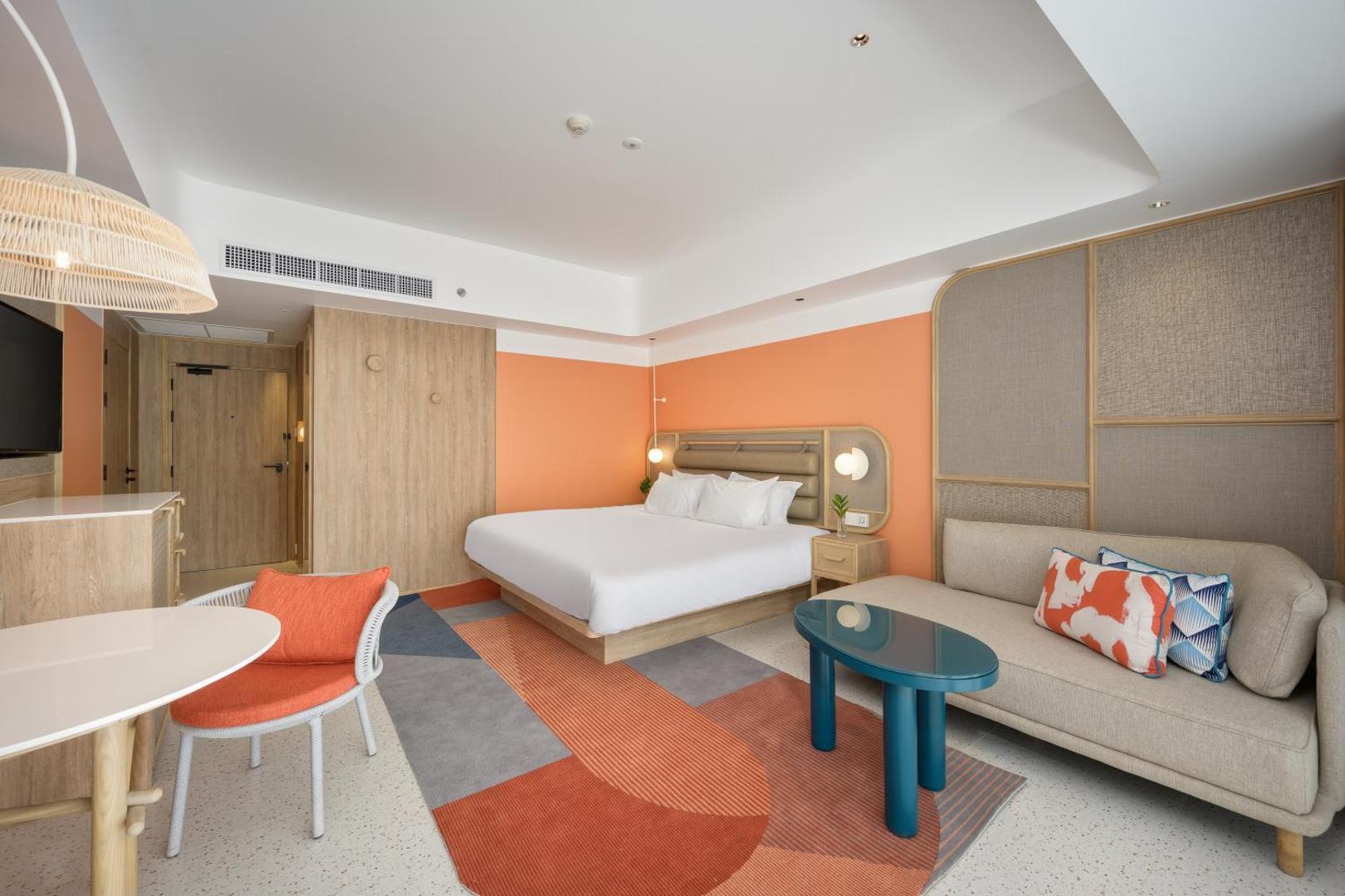 Centara Karon Resort Phuket moderns numurs ar oranžiem akcentiem, lielu gultu, dīvānu un stilīgu paklāju Puketas kūrortā, Taizemē