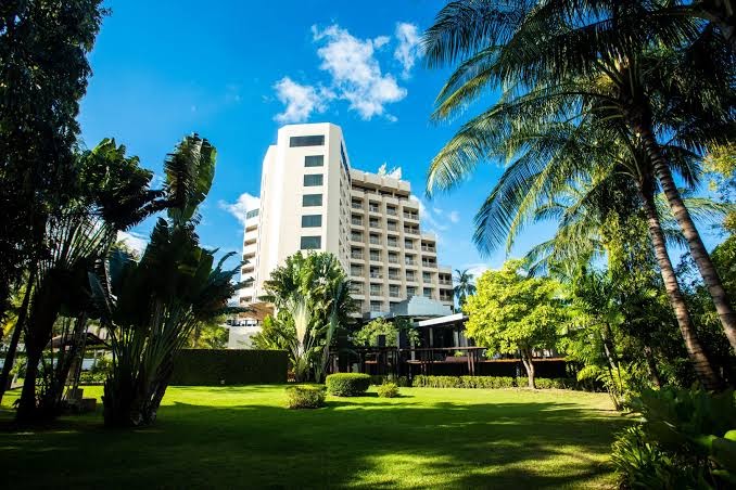 Centara Karon Resort Phuket daudzstāvu korpuss skatā no plaša, zaļa zālāja, ko ieskauj augstas palmas un tropu veģetācija