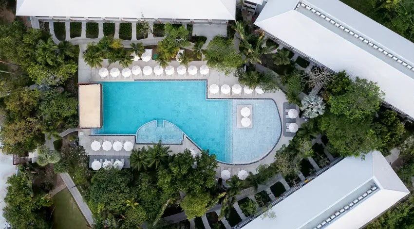 Aerofoto skats uz Centara Karon Resort Phuket galveno peldbaseinu, ko ieskauj balti saulessargi, palmas un viesnīcas korpusi