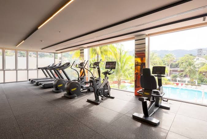 Centara Karon Resort Phuket moderns fitness centrs ar skatu uz baseinu un palmām — trenažieru zāle ar skrejceliņiem un velotrenažieriem