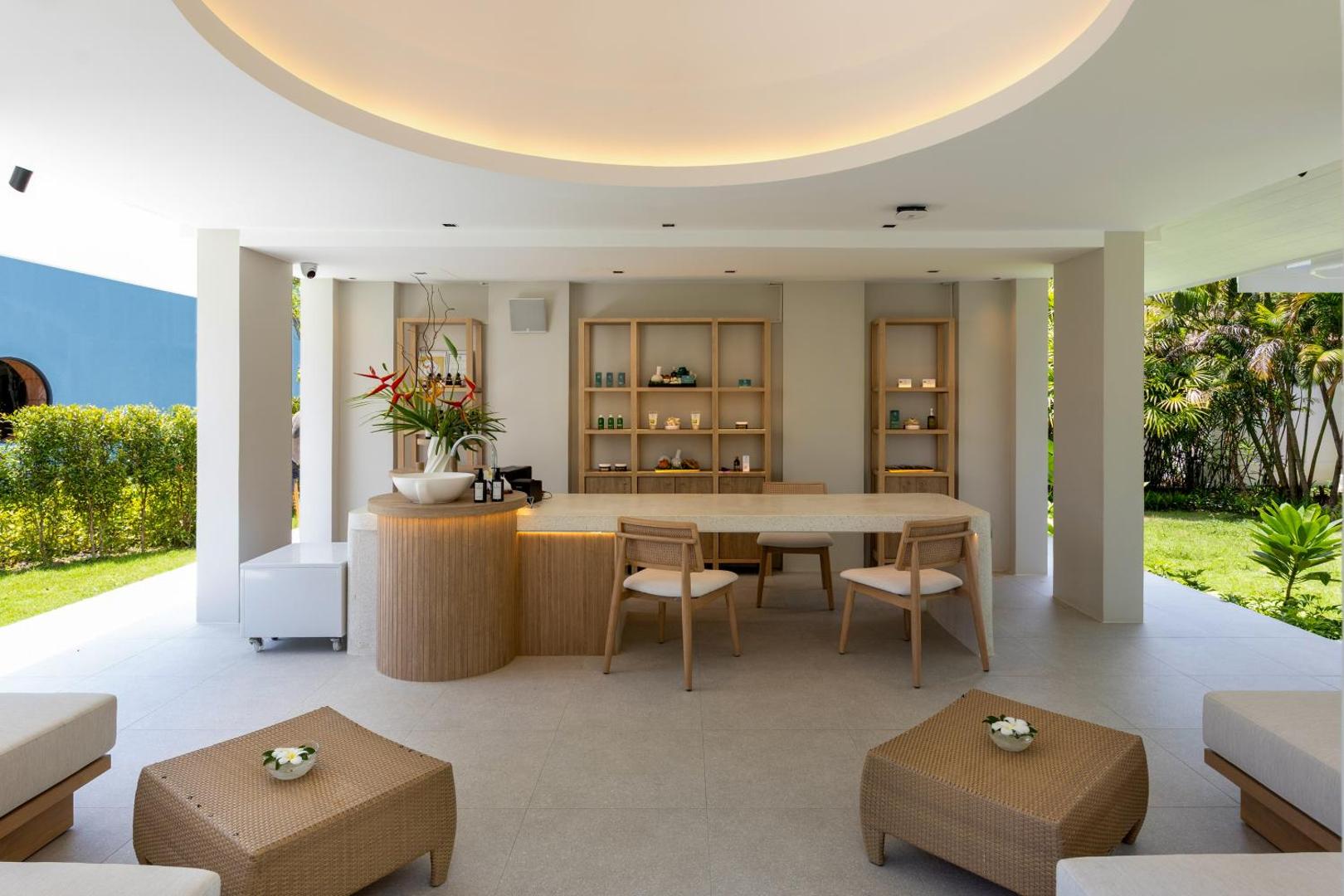 Centara Karon Resort Phuket spa centra recepcija ar gaišām koka mēbelēm, dabisku apgaismojumu un skatu uz tropisko dārzu