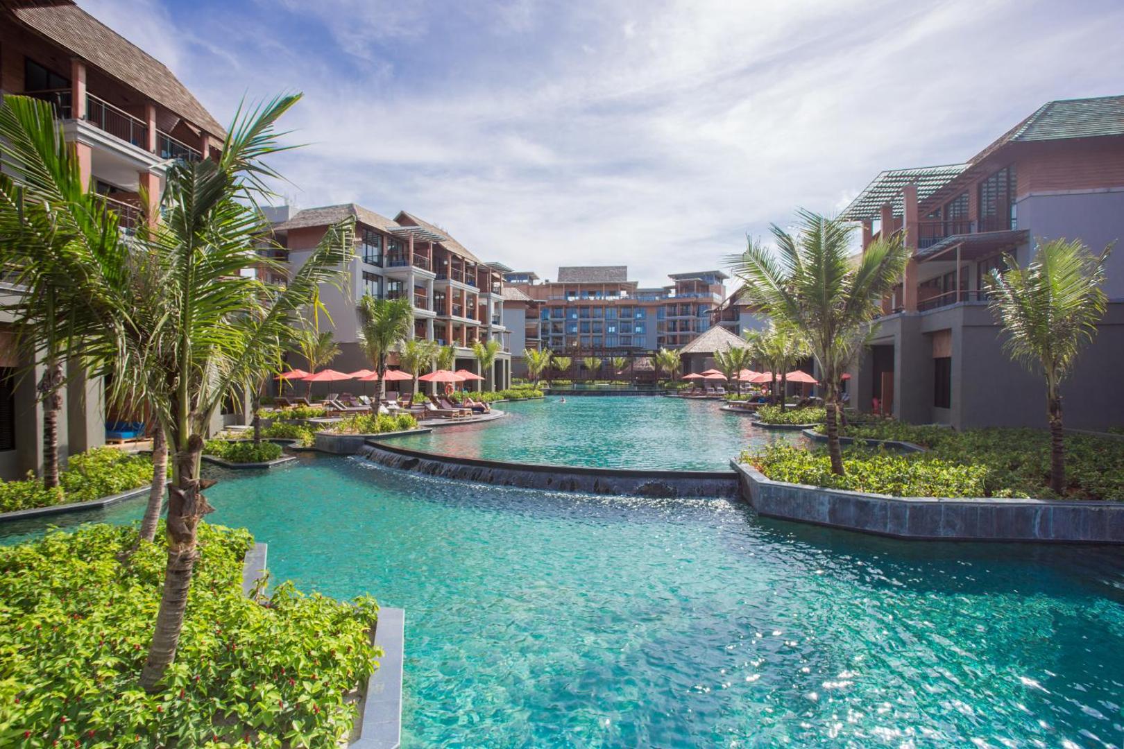 Mai Khao Lak Beach Resort & Spa plašais lagūnas tipa baseins starp viesnīcas korpusiem ar palmām un saulessargiem