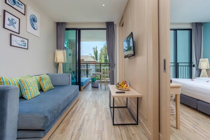 Mai Khao Lak Beach Resort gaišs viesnīcas numurs ar zilu dīvānu, koka grīdu un skatu uz balkonu un blakus esošo guļamistabu
