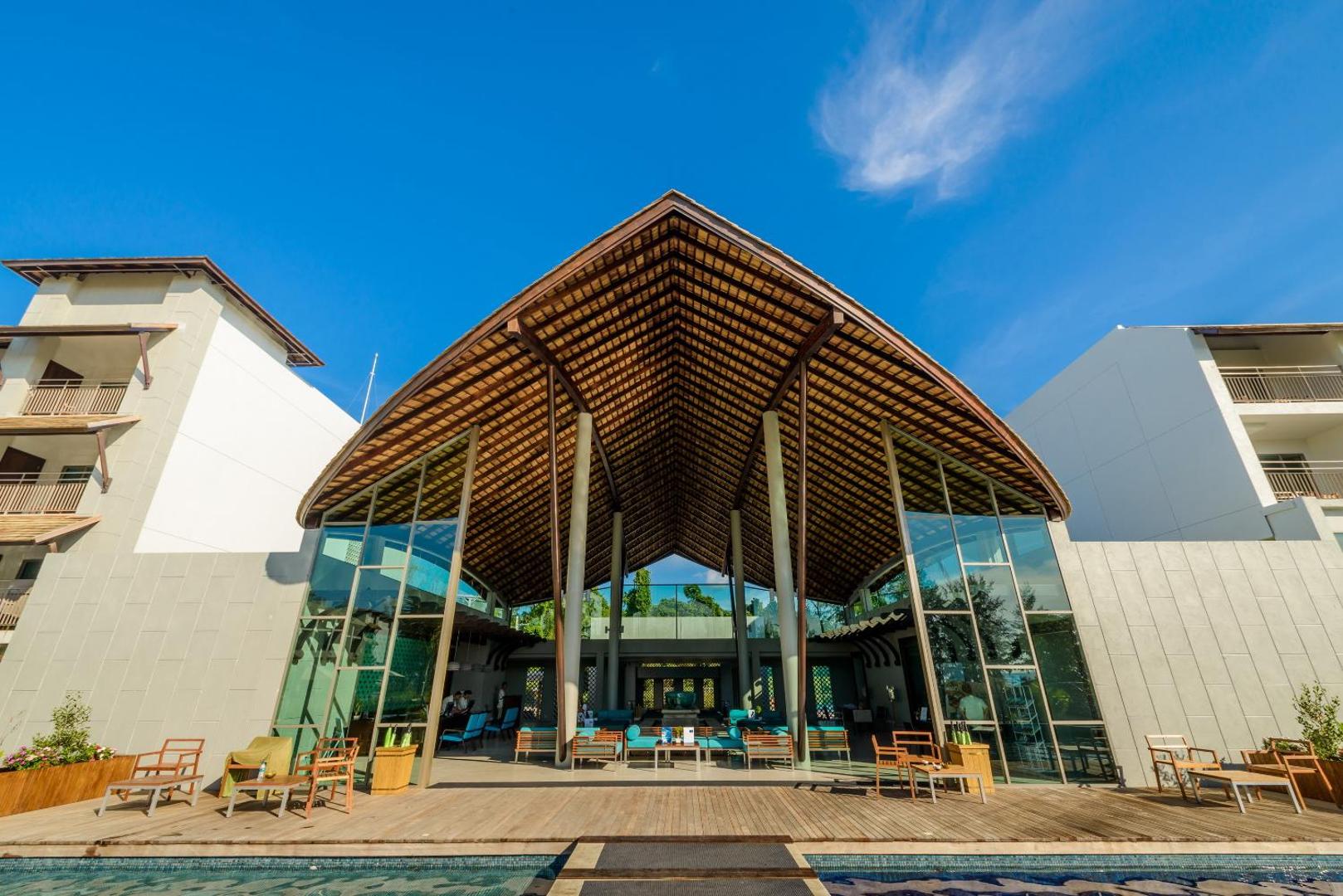Mai Khao Lak Beach Resort & Spa galvenā ēka ar iespaidīgu koka konstrukcijas jumtu un baseinu priekšplānā saulainā dienā