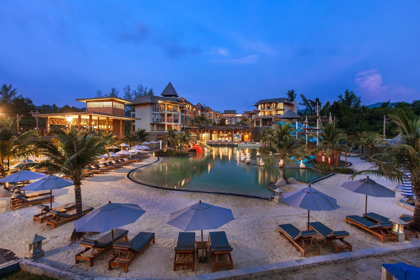 Mai Khao Lak Beach Resort & Spa centrālais baseins vakarā ar izgaismotām ēkām, sauļošanās krēsliem smiltīs un bērnu ūdens slidkalniņiem