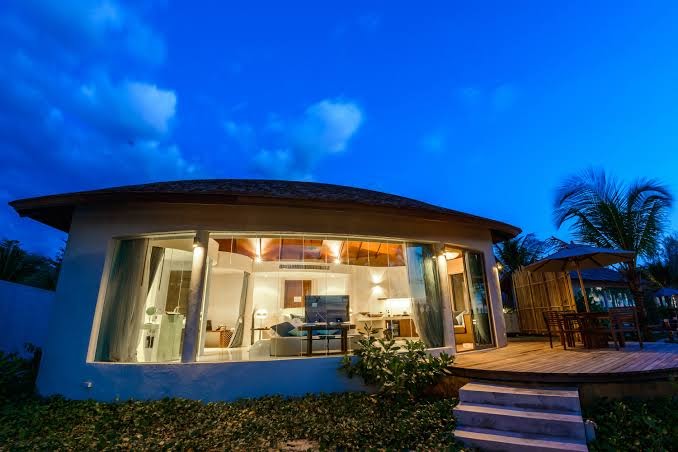 Mai Khao Lak Beach Resort moderna bungalo tipa ēka vakarā ar apgaismotu interjeru, koka terasi un dārza mēbelēm