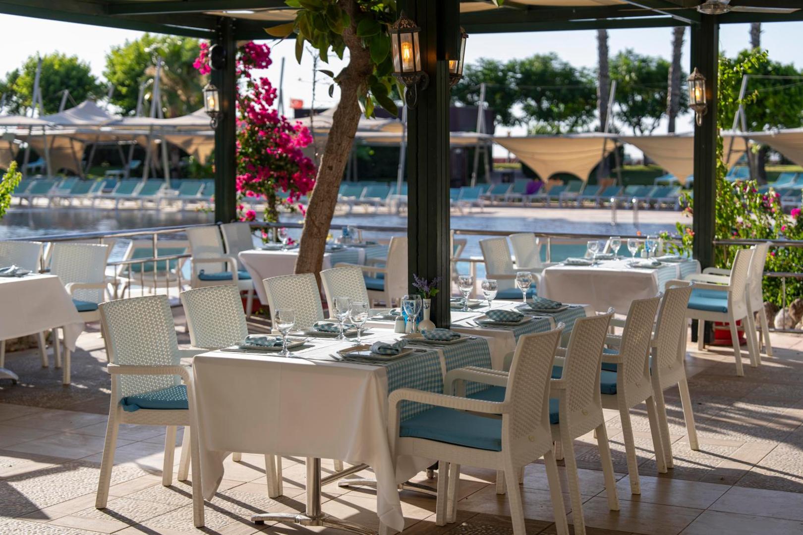 Limak Limra Hotel & Resort āra terases restorāns pie baseina ar balti klātiem galdiem un ziedošiem bugenvillu krūmiem.