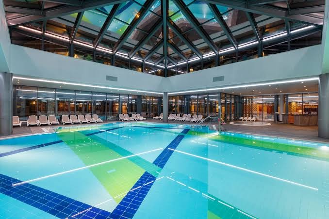 Limak Limra Hotel & Resort moderns iekštelpu baseins ar stikla jumtu un krāsainu flīžu dizainu — atpūtas krēsli pie logiem
