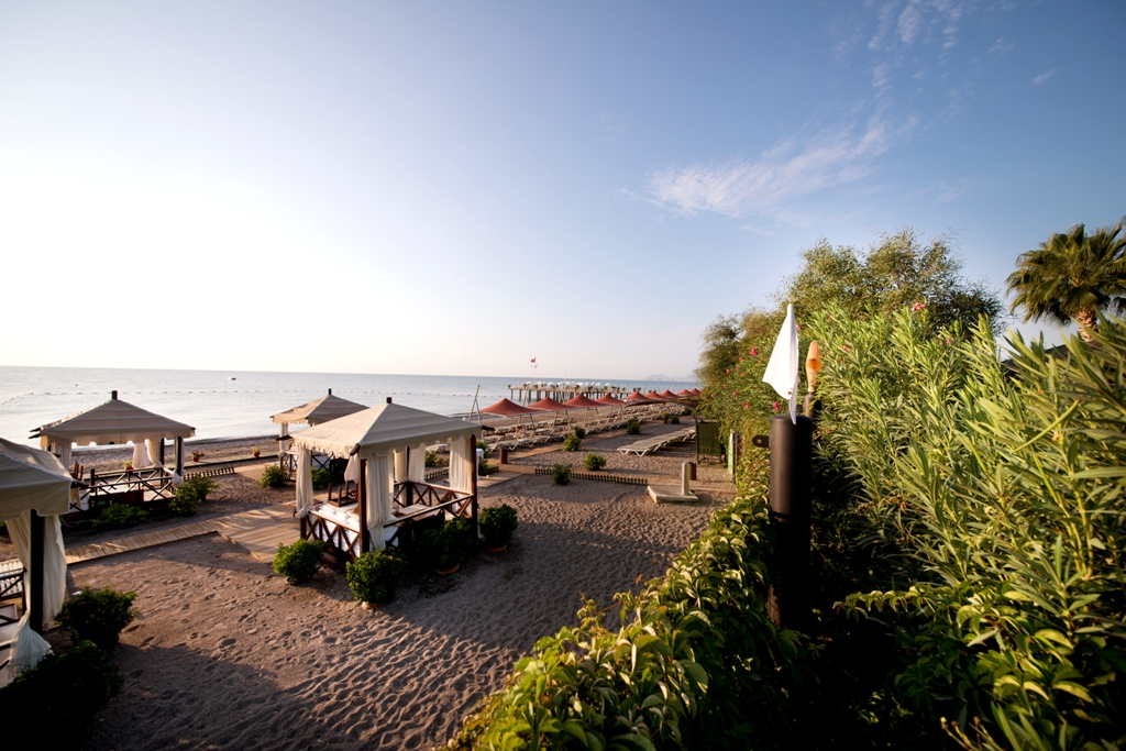 Limak Limra Hotel & Resort privātā pludmale saulrietā ar atpūtas teltīm, saulessargiem un koka laipām smiltīs