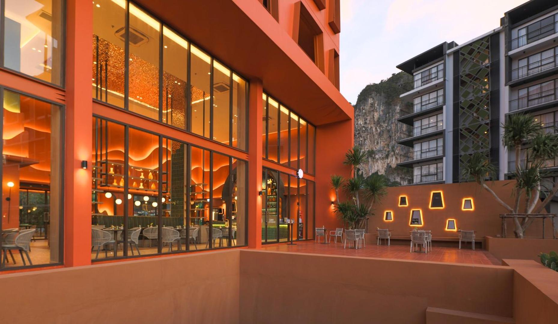Hotel Adam Krabi modernā arhitektūra ar oranžu fasādi un āra terasi — fonā redzamas iespaidīgās Krētas reģiona klintis un palmas