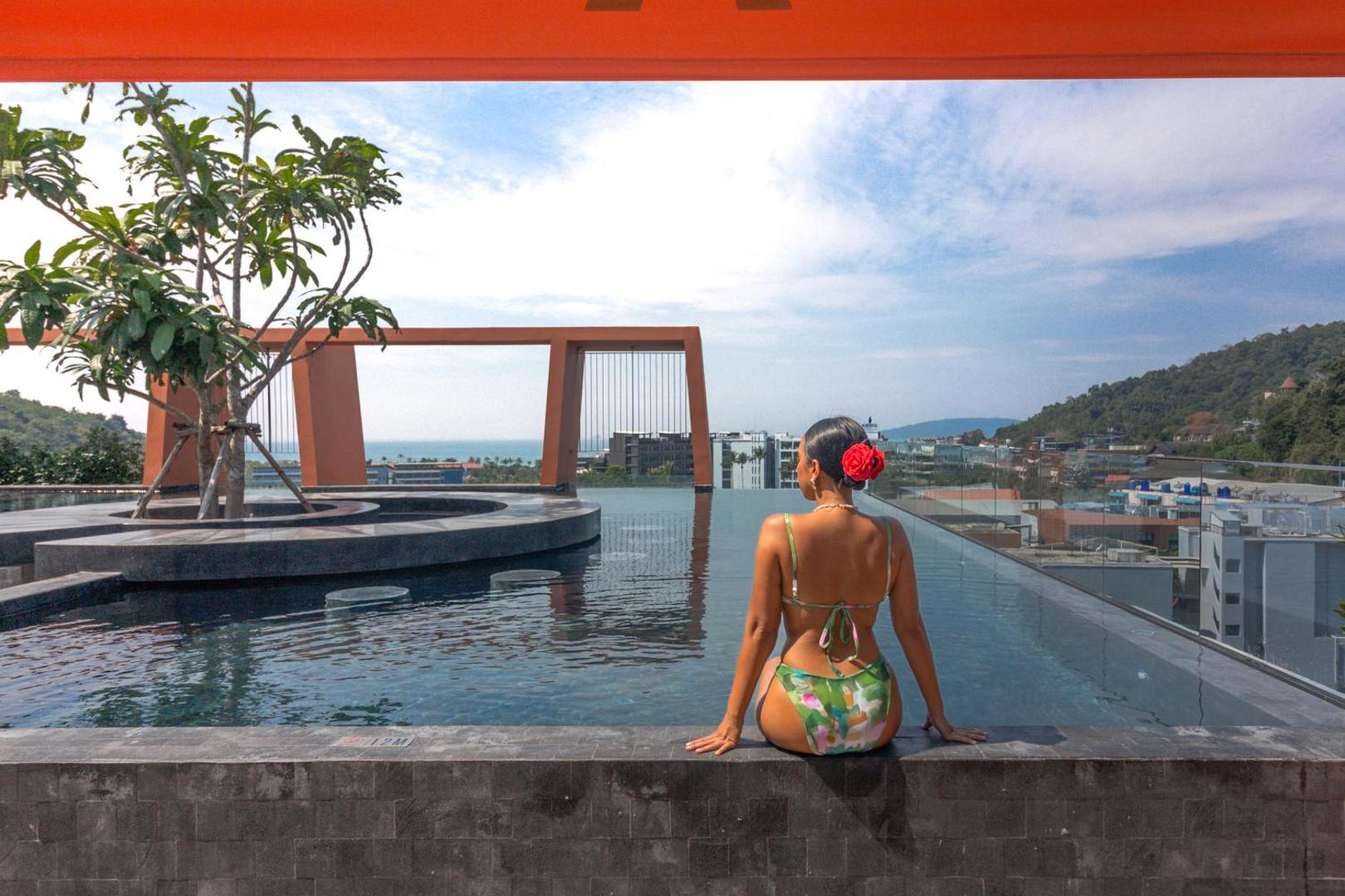 Sieviete pie Hotel Adam Krabi bezgalības baseina uz jumta — tālumā redzama jūra un pilsētas panorāma saulainā dienā