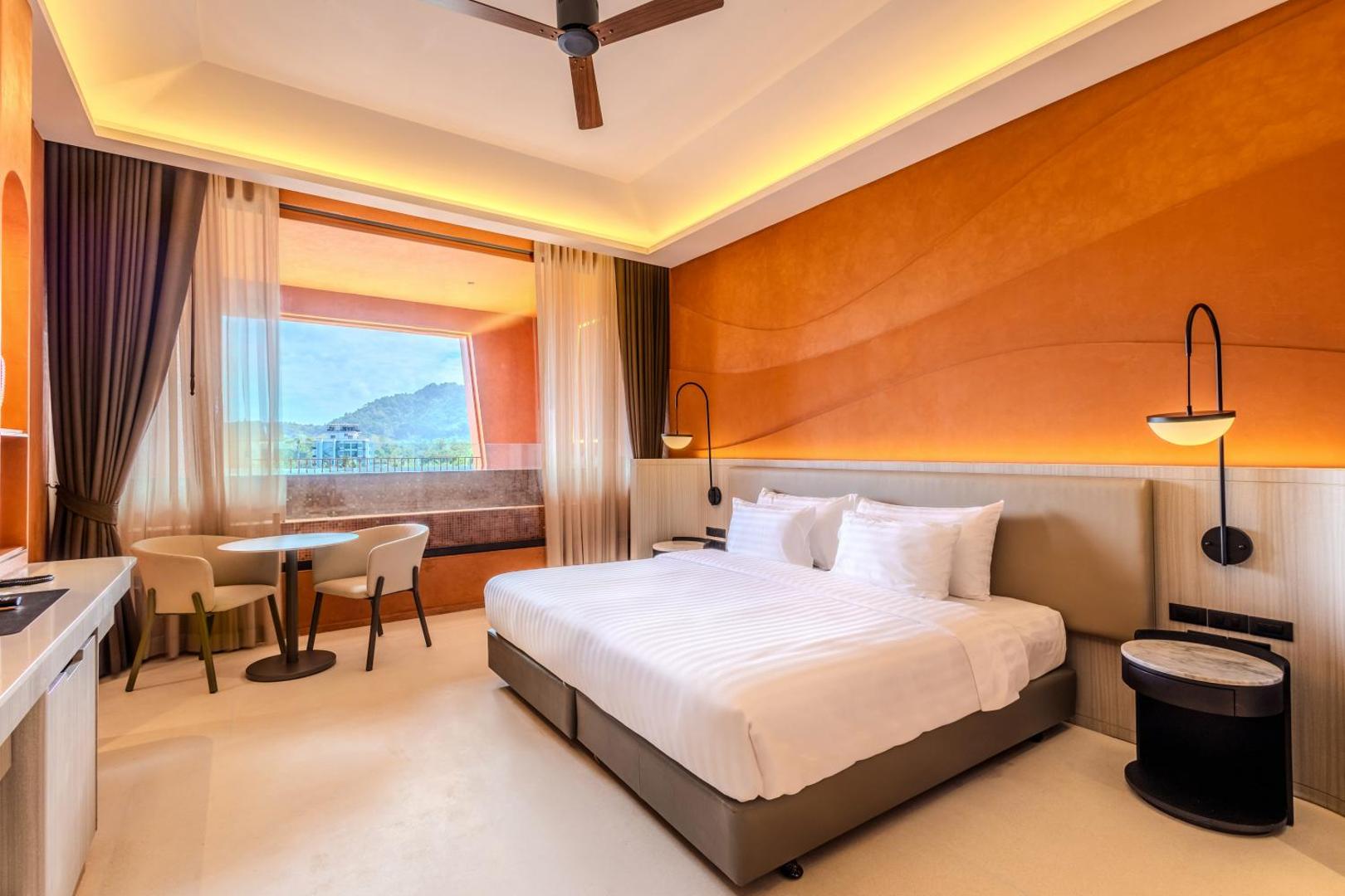 Gaišs Hotel Adam Krabi viesnīcas numurs ar lielu gultu, oranžu akcenta sienu un skatu uz kalniem tālumā
