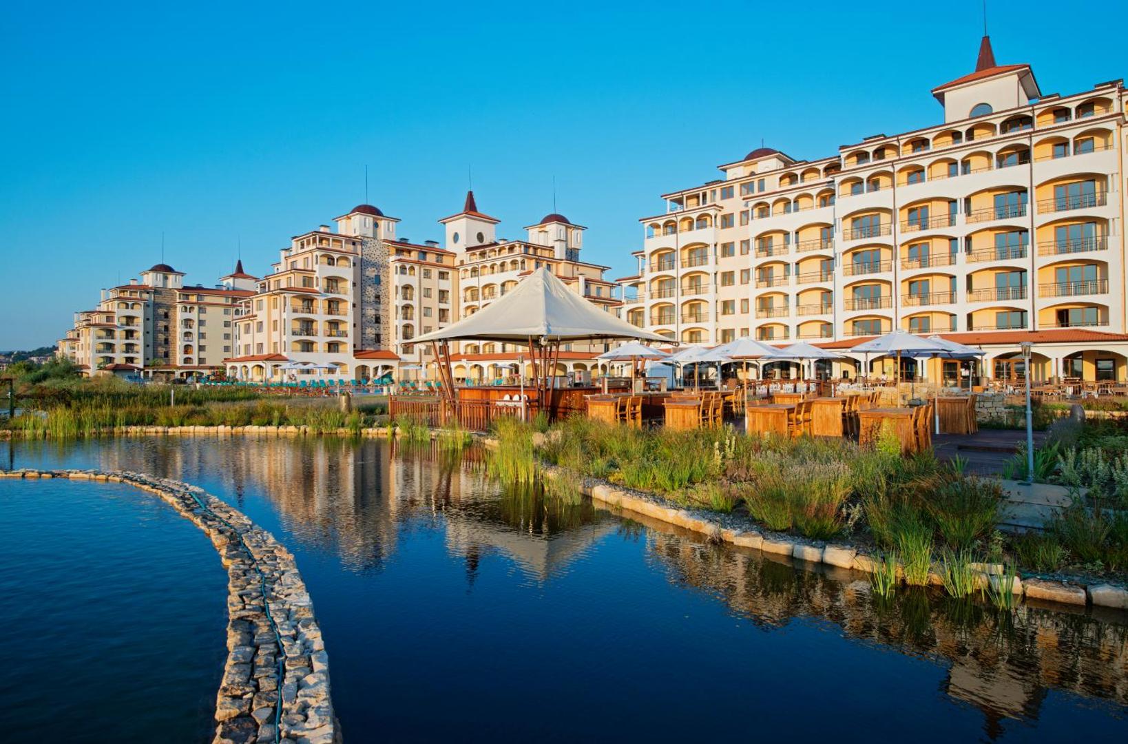 Sunrise All Suites Resort āra terase pie dīķa ar saulessargiem un galdiņiem, fonā redzama viesnīcas arhitektūra un zilas debesis